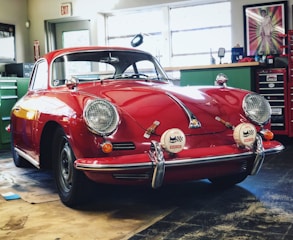 A classic red car with a vintage design is parked inside a garage or workshop. The car has a sleek and rounded body with chrome details and distinctive round headlights. Various tools and equipment are visible in the background, along with a tool chest and a framed picture on the wall.