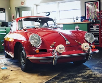 A classic red car with a vintage design is parked inside a garage or workshop. The car has a sleek and rounded body with chrome details and distinctive round headlights. Various tools and equipment are visible in the background, along with a tool chest and a framed picture on the wall.