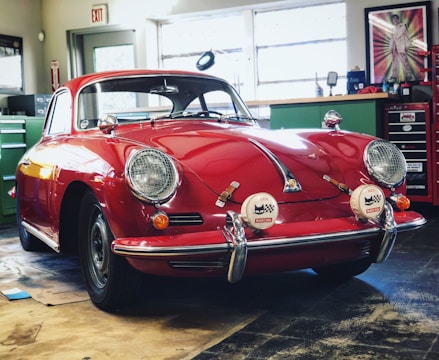 A classic red car with a vintage design is parked inside a garage or workshop. The car has a sleek and rounded body with chrome details and distinctive round headlights. Various tools and equipment are visible in the background, along with a tool chest and a framed picture on the wall.