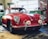 A classic red car with a vintage design is parked inside a garage or workshop. The car has a sleek and rounded body with chrome details and distinctive round headlights. Various tools and equipment are visible in the background, along with a tool chest and a framed picture on the wall.