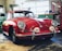 A classic red car with a vintage design is parked inside a garage or workshop. The car has a sleek and rounded body with chrome details and distinctive round headlights. Various tools and equipment are visible in the background, along with a tool chest and a framed picture on the wall.