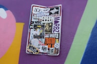 A weathered street sign is covered in a variety of stickers, ranging from colorful designs to monochrome logos and text. The background is a vibrant wall painted with purple, pink, and blue hues.