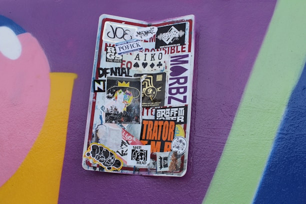 A weathered street sign is covered in a variety of stickers, ranging from colorful designs to monochrome logos and text. The background is a vibrant wall painted with purple, pink, and blue hues.