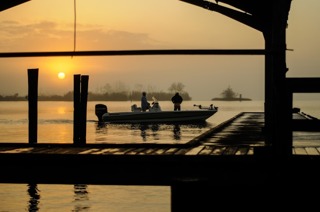 A serene lakeside scene at sunset, with a small boat in the water. Two individuals are on the boat, silhouetted against the orange and yellow sky. The sun is low on the horizon, casting a warm glow over the water. A wooden structure partially frames the view, adding depth to the image.