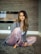 A woman with long hair sits comfortably on a soft gray rug in a room with wooden panel walls. She is wearing a gray cardigan over a white top with lace details and pink satin pants. Her expression is serene, and she is barefoot.