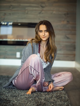 A woman with long hair sits comfortably on a soft gray rug in a room with wooden panel walls. She is wearing a gray cardigan over a white top with lace details and pink satin pants. Her expression is serene, and she is barefoot.