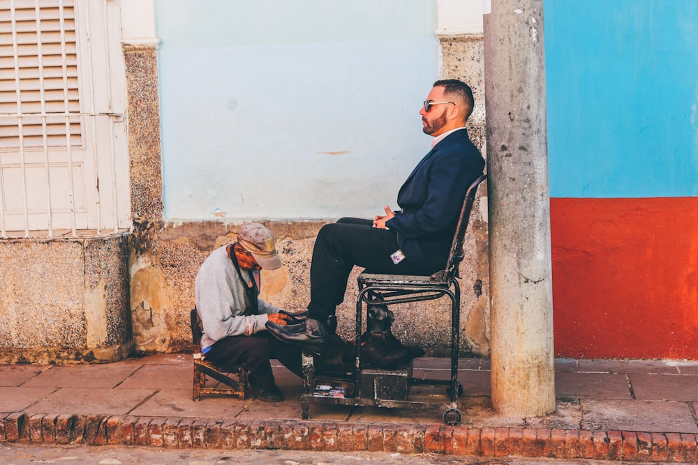 man cleaning shoe of another man near white painted wall outdoors