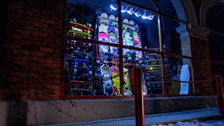 A nighttime window display is filled with a colorful array of skateboards stacked vertically. There are neon lights and various graphic designs on the boards, including vibrant patterns and text. A chain is visible in front of the shop, and brick walls frame the left side.