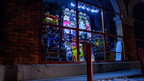 A nighttime window display is filled with a colorful array of skateboards stacked vertically. There are neon lights and various graphic designs on the boards, including vibrant patterns and text. A chain is visible in front of the shop, and brick walls frame the left side.