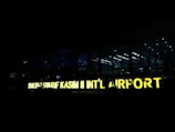 A large, illuminated sign displays the name 'SULTAN SYARIF KASIM II INT’L AIRPORT' against a backdrop of a modern airport terminal at night. The terminal is made of glass, allowing interior lights to shine through, creating a contrast with the dark surroundings.