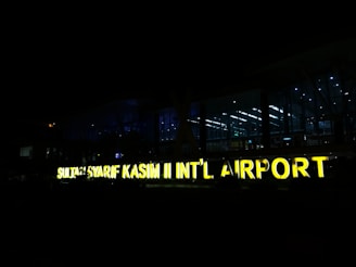 A large, illuminated sign displays the name 'SULTAN SYARIF KASIM II INT’L AIRPORT' against a backdrop of a modern airport terminal at night. The terminal is made of glass, allowing interior lights to shine through, creating a contrast with the dark surroundings.