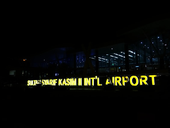 A large, illuminated sign displays the name 'SULTAN SYARIF KASIM II INT’L AIRPORT' against a backdrop of a modern airport terminal at night. The terminal is made of glass, allowing interior lights to shine through, creating a contrast with the dark surroundings.