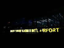 A large, illuminated sign displays the name 'SULTAN SYARIF KASIM II INT&rsquo;L AIRPORT' against a backdrop of a modern airport terminal at night. The terminal is made of glass, allowing interior lights to shine through, creating a contrast with the dark surroundings.