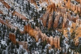 A rugged landscape with towering rock formations known as hoodoos, interspersed with patches of snow and green coniferous trees. The scene is set against a backdrop of reddish-orange and brown rock layers, creating a striking natural contrast.