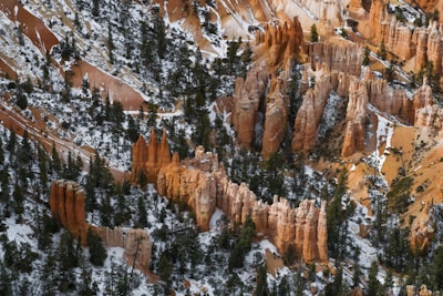 A rugged landscape with towering rock formations known as hoodoos, interspersed with patches of snow and green coniferous trees. The scene is set against a backdrop of reddish-orange and brown rock layers, creating a striking natural contrast.
