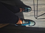 A pair of legs in black leggings are visible, with feet wearing blue and black athletic sneakers. The feet are positioned on a wooden gray deck with visible grain patterns. There is a metal chair leg casting a shadow on the deck.