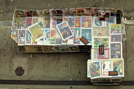 A sidewalk display consisting of an array of vintage movie and concert posters. The posters are neatly arranged in a rectangular shape on the ground, featuring iconic films and events. A variety of vibrant, colorful designs are visible, with some posters featuring notable classic movies and musical legends.