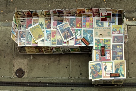 A sidewalk display consisting of an array of vintage movie and concert posters. The posters are neatly arranged in a rectangular shape on the ground, featuring iconic films and events. A variety of vibrant, colorful designs are visible, with some posters featuring notable classic movies and musical legends.