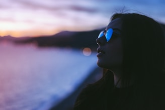 A silhouette of a person wearing sunglasses against a backdrop of a serene evening landscape, featuring a soft focus on a distant shoreline with gentle waves and a colorful sky at dusk.