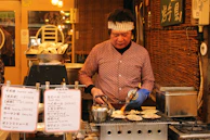 A close-up of a street food vendor preparing takoyaki, steam rising from the hot octopus balls.