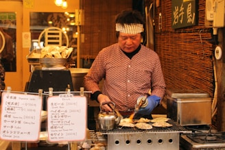 A street vendor is preparing seafood on a grill, using a blowtorch to cook the food. The vendor is wearing a patterned shirt and a headband, standing in front of a wooden stall. Several signs with Japanese writing and menus featuring drinks and food prices are placed at the front of the stall. A tray filled with oysters sits nearby.