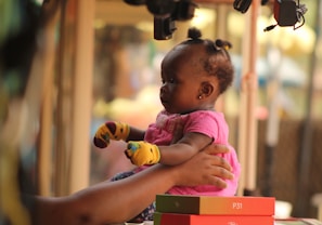 A baby wearing a pink outfit and colorful mittens is being held gently by a pair of adult hands. The setting appears to be indoors, possibly in a market or a street vendor stall, judging by the background with blurred items and lighting.