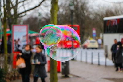 A large soap bubble with iridescent colors floats in an urban setting. People in the background are blurred, perhaps indicating movement, and red buses and trees are visible, suggesting a city environment.