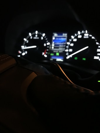 The image depicts a close-up of a car dashboard at night, featuring illuminated gauges and dials. The focus is on the brightly lit speedometer and tachometer, with glowing indicators and a small screen displaying information. The steering wheel is partially visible, with shadows and reflections adding depth.
