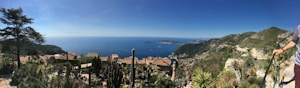 A panoramic view of a coastal landscape featuring vivid blue sea and clear skies. The foreground contains a variety of cacti and succulents, surrounded by traditional terracotta-roofed buildings. Rolling hills and mountains stretch towards the horizon, providing a sense of vastness and serenity.