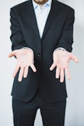 A person in a dark suit is standing with their arms extended forward, palms facing up in a gesture of openness or offering. The background is plain and light-colored, creating a sharp contrast with the dark suit.