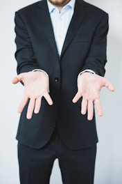 A person in a dark suit is standing with their arms extended forward, palms facing up in a gesture of openness or offering. The background is plain and light-colored, creating a sharp contrast with the dark suit.