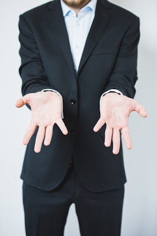 A person in a dark suit is standing with their arms extended forward, palms facing up in a gesture of openness or offering. The background is plain and light-colored, creating a sharp contrast with the dark suit.
