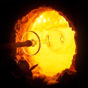 A glassblowing furnace glows intensely with a bright orange and yellow light. A glass object, resembling a wine glass, is being formed on a blowpipe as it is inserted into the furnace. The contrasting darkness around the furnace opening highlights the radiant heat and light emanating from within, creating a dramatic and vivid scene.