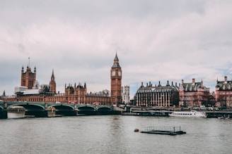 A panoramic view of the iconic Houses of Parliament along the River Thames. The famous clock tower stands prominently, while a bridge crosses the water. A few boats can be seen on the river, with a cloudy sky overhead.