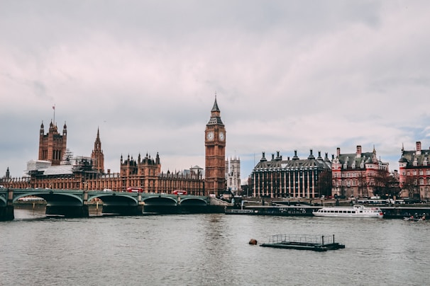 A panoramic view of the iconic Houses of Parliament along the River Thames. The famous clock tower stands prominently, while a bridge crosses the water. A few boats can be seen on the river, with a cloudy sky overhead.