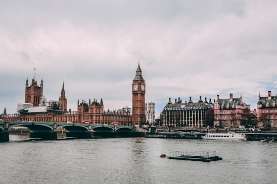 A panoramic view of the iconic Houses of Parliament along the River Thames. The famous clock tower stands prominently, while a bridge crosses the water. A few boats can be seen on the river, with a cloudy sky overhead.