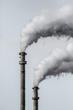 Two industrial smokestacks are emitting large plumes of white smoke against a pale gray sky. The details of the smokestacks' metallic structure are visible, and the smoke is billowing upwards, suggesting active emissions.
