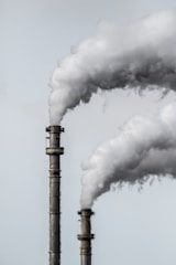 Two industrial smokestacks are emitting large plumes of white smoke against a pale gray sky. The details of the smokestacks' metallic structure are visible, and the smoke is billowing upwards, suggesting active emissions.