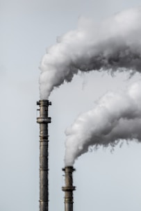 Two industrial smokestacks are emitting large plumes of white smoke against a pale gray sky. The details of the smokestacks' metallic structure are visible, and the smoke is billowing upwards, suggesting active emissions.