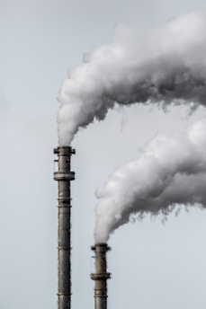 Two industrial smokestacks are emitting large plumes of white smoke against a pale gray sky. The details of the smokestacks' metallic structure are visible, and the smoke is billowing upwards, suggesting active emissions.