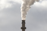 A large industrial smokestack emits a dense plume of white smoke into a cloudy sky, indicating active combustion or industrial activity.