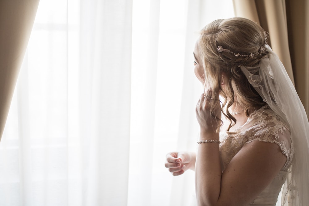 Mujer con vestido de novia blanco cerca de la cortina de la ventana