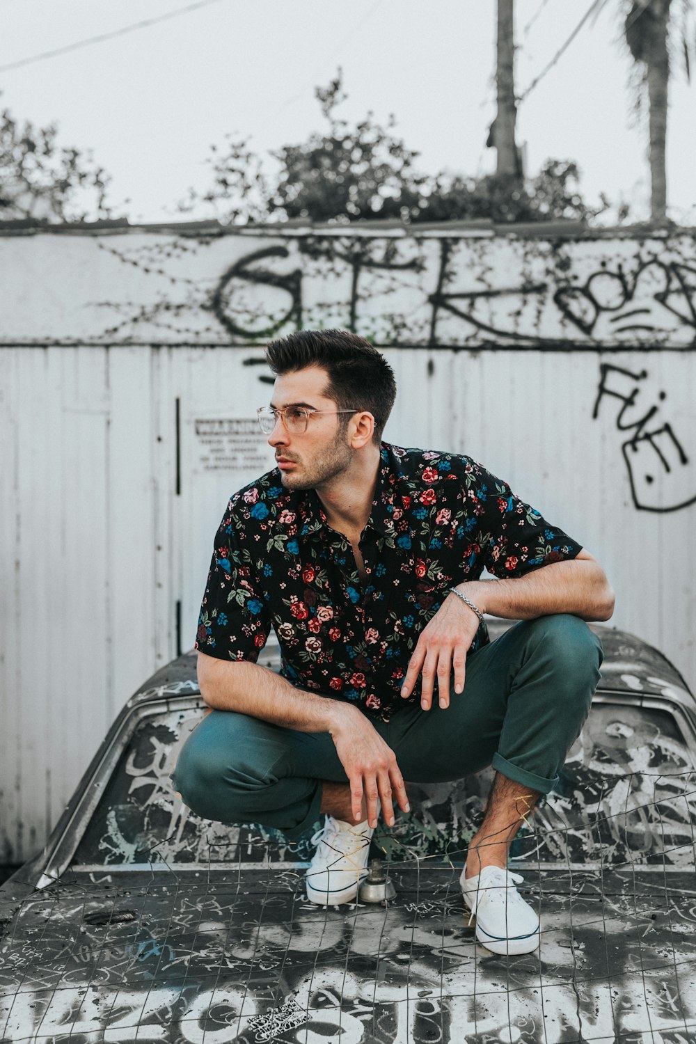 uomo in camicia con colletto abbottonato floreale nero, blu e rosso e pantaloni grigi seduto sul cofano dell'auto durante il giorno