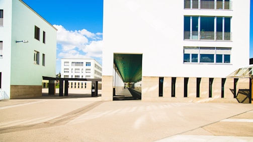 Modern architectural buildings with a clean design are set against a clear blue sky. The structures feature large windows and light-colored walls, creating a bright and open atmosphere. An open walkway between the buildings adds depth to the scene.