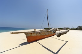 A traditional wooden boat rests on a pristine sandy beach with a clear blue sky above. The ocean water nearby is a rich blue, stretching towards the horizon. The beach appears deserted and peaceful, with some vegetation visible in the distance.