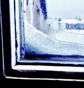 A window with a significant accumulation of snow and ice on the outside. The window frame displays peeling paint, suggesting wear and exposure to cold weather. In the background, buildings and a snow-covered landscape are visible, hinting at a cold and wintry environment.
