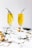 Two champagne flutes containing a golden-yellow beverage with sprigs of thyme. The glasses are placed on a white surface adorned with gold ornaments and green sprigs, creating a festive and elegant setting.