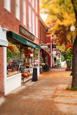 A quaint, small-town street lined with charming brick buildings, some of which have awnings and shop displays. The sidewalk is decorated with pumpkins and potted flowers, and the scene is framed by trees showcasing vibrant autumn foliage.