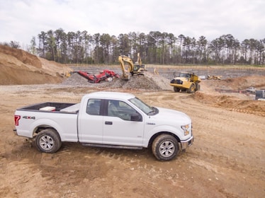 A white pickup truck is parked on a dirt construction site with machinery in the background, including an excavator and a dump truck. The area is surrounded by mounds of dirt and gravel, with a line of trees visible in the distance under a cloudy sky.