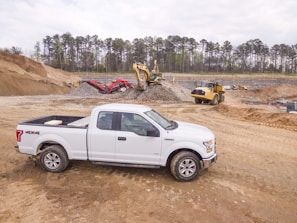A white pickup truck is parked on a dirt construction site with machinery in the background, including an excavator and a dump truck. The area is surrounded by mounds of dirt and gravel, with a line of trees visible in the distance under a cloudy sky.